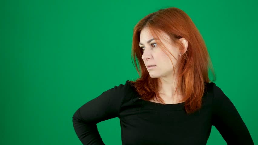 Dissatisfaction, lips pursed, hands at sides, woman in profile, eyes lowered red hair young woman on a green background in a chroma key studio emotions close-up brown eyes Light skin European
