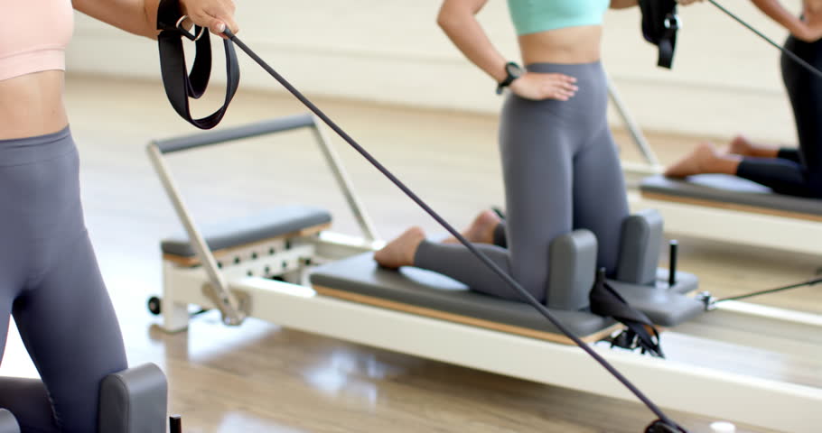 Two women engage in a Pilates workout using reformer machines. The biracial woman in the foreground wears a teal sports bra and grey leggings, while her counterpart dons black.