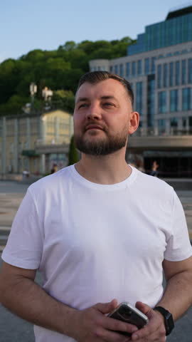 Bearded man in white t-shirt holding smartphone, standing in urban square with historical buildings in background, on clear day