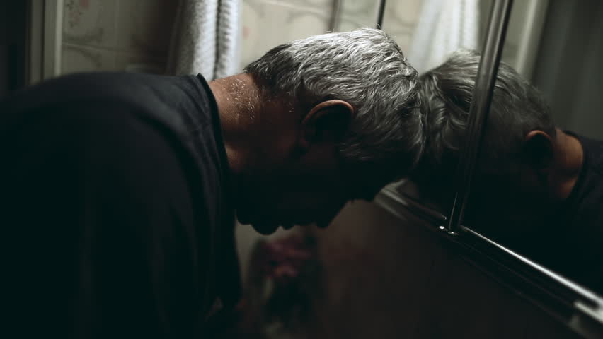 Depressed burned senior man gazing at his own mirror reflection standing in bathroom by sink feeling despair during emotional crisis, elderly person in desperation