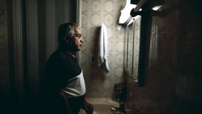 Tired senior man looking at his own reflection standing in bathroom mirror, profile body in humble residence depicting old age - Powered by Shutterstock - Get 15% off with code: PIKWIZARD15
