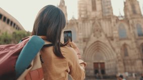 Traveler girl, with backpack on her shoulders, takes photo on mobile phone of an old building in the historical part of European city, back view
