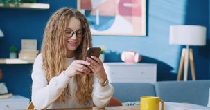 Portrait of cheerful female holding smartphone. Young woman sitting at table. Female relaxing at stylish apartment. Pretty woman texting by smartphone. Female using new new. Online communication.