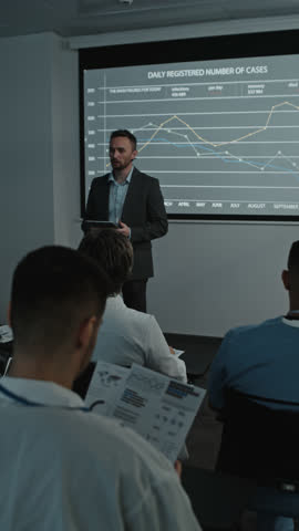 Vertical shot of male speaker in formal suit holding digital tablet and commenting on presentation on projected screen in front of audience during medical conference