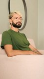 Vertical video, Young blond man with a beard wearing an olive green T-shirt relaxes while sitting on the sofa in a cozy living room