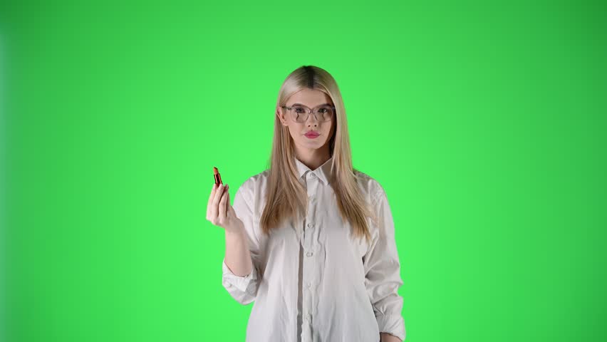 Woman with white shirt and glasses puts on red lipstick, green background