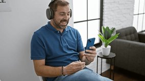 Handsome middle-aged man with grey hair video calling on smartphone indoors, wearing headphones, seated in a modern lobby.