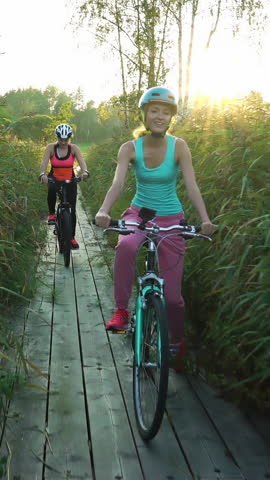Two young women ride bicycles on a wooden ecological trail among the reeds. Vertical video