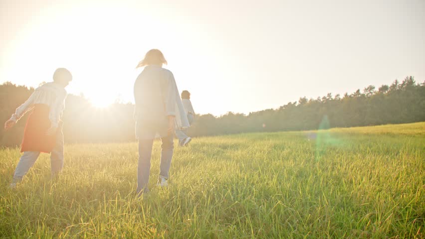 Camera view from behind of three women walking up hill with green grass. Sun shining very bright. People living actively. Females walking and jumping together through wide field and smiling.