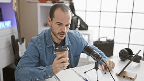 A bald man with a beard sips coffee while podcasting indoors using a microphone and tablet.