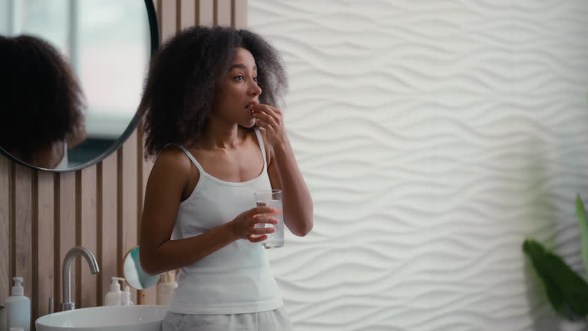 Healthy African American beautiful woman sick ill girl female taking pill with glass of water drinking remedy drug vitamins supplements at morning bathroom daily medication for beauty skin hair care