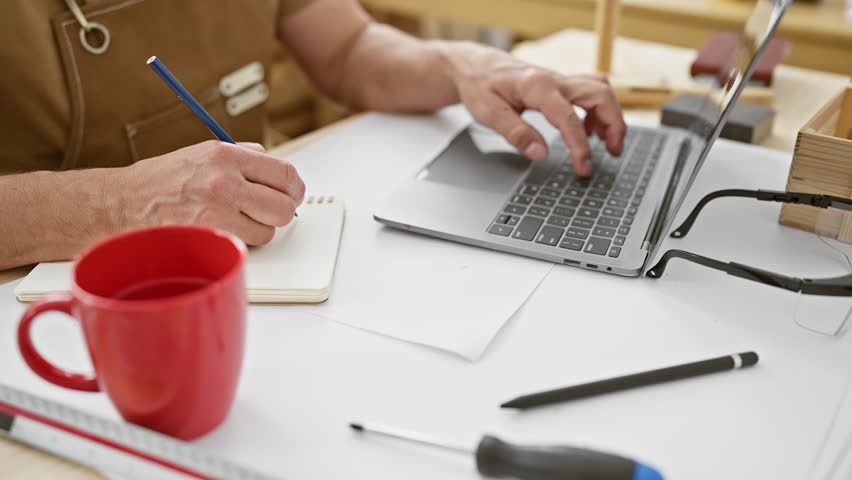A mature man sketches a design in a carpentry workshop beside a laptop and coffee mug.