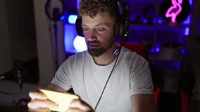 A young, bearded, handsome man with blue eyes wearing headphones waves while gaming in a dark indoor room.