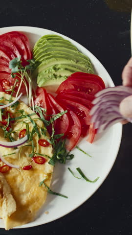 Vertical top down shot of hand of chef adding red onion slices to omelet served with vegetables on plate