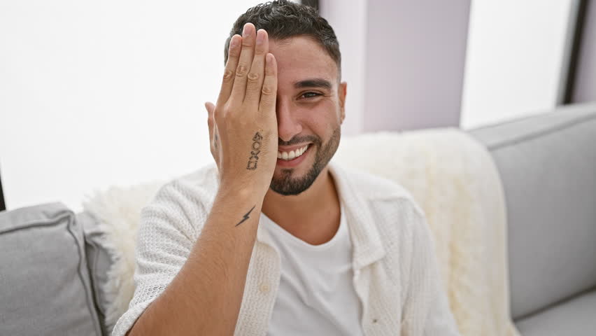 At home, a confident young arab man sitting on his living room sofa, covering one eye with his hand, a cheerful smile gracing his face, radiates a surprising sense of fun.