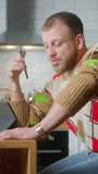 Vertical video. Man sitting on chair in kitchen with tablet on table, talking on speakerphone on his mobile phone. Male relaxing at home, communicating with family and friends. Side shot