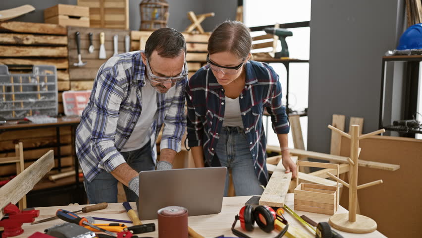 Two professional carpenters engaged in a serious conversation about their woodwork project, using a laptop to navigate through their carpentry blueprint while holding a timber plank indoors