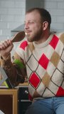 Vertical video. Cosy adult man singing into a ladle like a microphone while sit in the kitchen at home