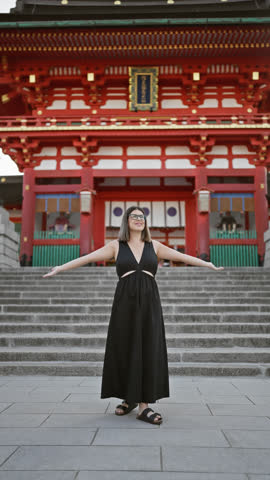 Joyful hispanic woman in glasses, embracing freedom with open arms, smiling and looking around fushimi inari taisha shrine in kyoto, japan
