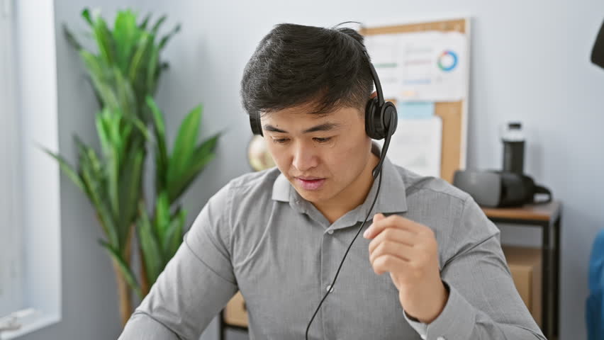A focused asian man wearing a headset interacts with technology in a modern office setting. Royalty-Free Stock Footage #3459407803
