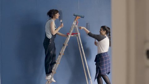 Two happy smiling girls  have fun dancing, singing and painting the blue wall in living room.  