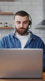 Vertical video. Bearded young adult business man wear wireless headset sitting against the kitchen counter having video conference calling on laptop computer talk by webcam in online chat