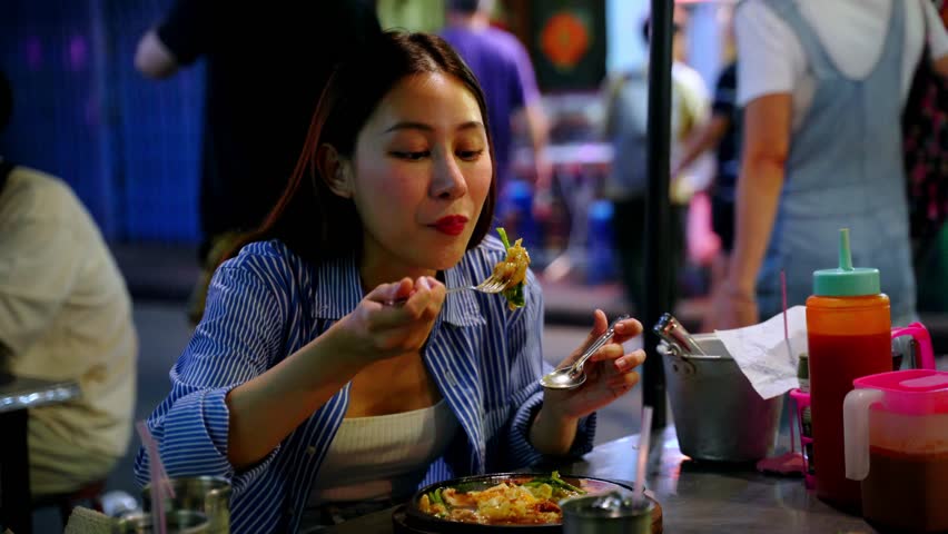 Young adult Asian woman enjoys eating a local Thai food in the street food stall shop at night in Bangkok, Thailand. Solo backpacker eating street food on table in the street. Royalty-Free Stock Footage #3459615295