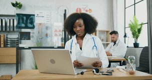 Woman doctor in white uniform making notes into papers during video meeting on laptop in modern medical office