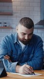 Vertical video. Bearded young adult man sitting against the kitchen counter working on tablet device taking notes on paper. Peaceful relaxing working