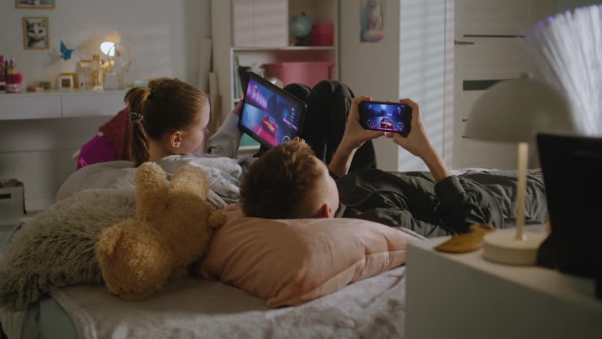 Two teenagers lie on bed in their room and play video games on digital devices. Girl uses tablet, boy uses phone to play together. Sister spending leisure time with her brother. Family relationship.