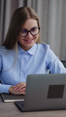 Positive young female employee looking through the information on her computer. Lady is typing on the laptop keyboard smiling. Office indoors backdrop. Vertical video. Royalty-Free Stock Footage #3459720977