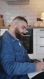 Vertical video. Young man student sitting on the floor leaning against the kitchen counter doing research working on laptop taking notes on paper while listening the music with in ear headphone