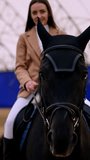 Positive smiling Caucasian lady in beige jacket riding a black horse. Low angle perspective. Blurred drill-hall at backdrop. Vertical video.