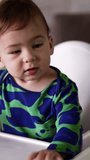 Lovely active baby boy in green spotted shirt sits at the dining table. Little kid waving hands and then stares at something. Close up. Vertical video.