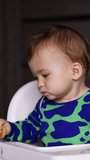Adorable baby boy with smudged face sitting in a chair. Baby holding a bagel and banging it by the table. Blurred backdrop. Vertical video.