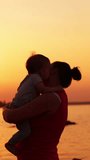 Brunette woman in red sleeveless shirt holding a little toddler. Mother and son enjoying beautiful sunset near the river. Vertical video