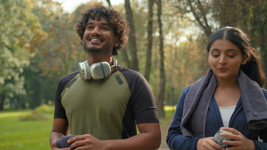 Happy smiling sport couple smile laughing talking walking together in park city after running workout talk going outdoors fitness instructor with client walk friendly Indian woman girl Arabian man guy