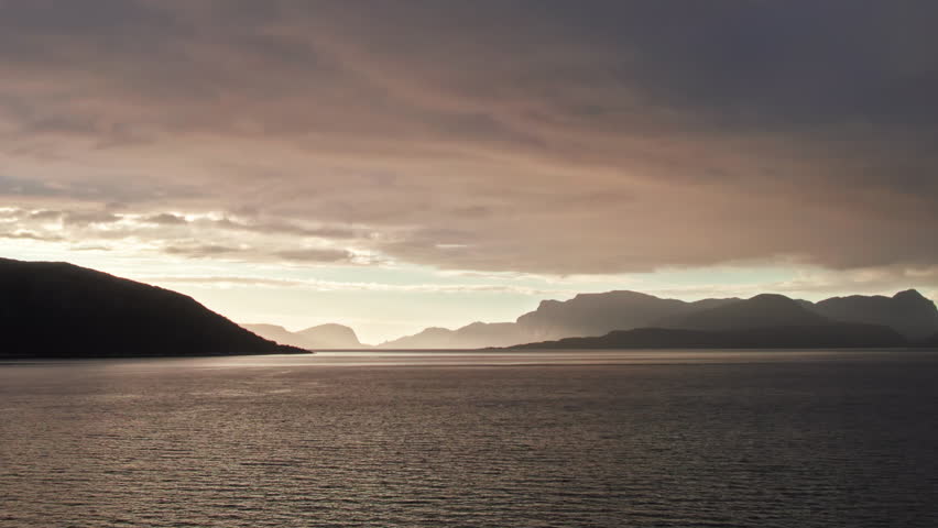 Panoramic aerial shot, sweeping over Sognefjord in Norway towards a bright sunset over the mountainous horizon. The sky is dark and cloudy. Royalty-Free Stock Footage #3459766405