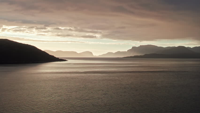 Aerial shot, climbing over the gently rippling water of Sognefjord in Norway, looking out at a sunset over multiple shadowy mountains. The sky is dark and overcast. Royalty-Free Stock Footage #3459766553