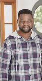 Vertical video of happy african american man with beard standing and smiling in sunny house. Lifestyle,relaxation,free time and domestic life, unaltered.