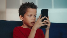 A little boy engages in an online game on phone, comfortably resting on a couch