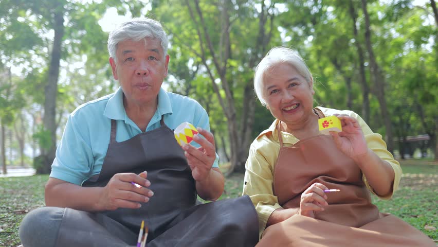 An elderly Asian couple does art activity painting ceramic pots in the garden outside. Living happily in retirement. art therapy