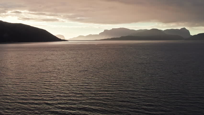 Aerial shot, sweeping over the rippling still water of Sognefjord in Norway, looking towards a sunset over shadowy mountainous islands in the distance. The sky is dark and moody. Royalty-Free Stock Footage #3459923831