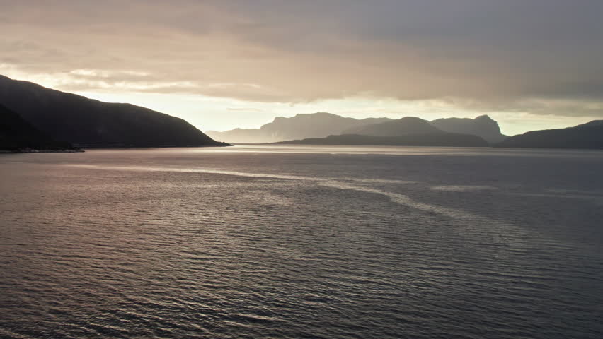 Aerial view, climbing over Sognefjord in Norway, moving toward a sunset over distant shadowy mountains and islands. The sky is dark and cloudy. Royalty-Free Stock Footage #3459923949