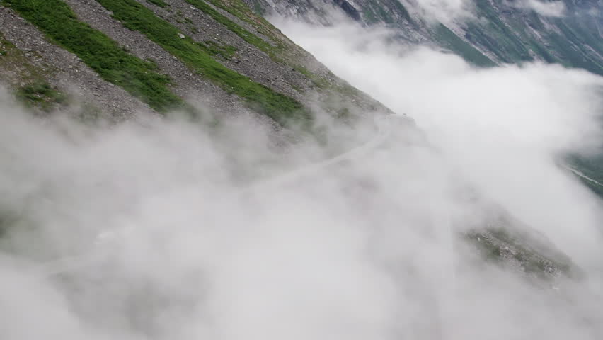 Aerial shot circling around a cloud covered hairpin bend on the Trollstigen in Norway as it snakes down the steep valley side. A Tour bus approaches the sharp turn. 