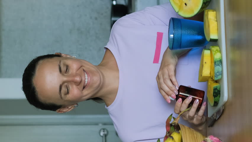 Vertical video. Young female surrounded by exotic tropical organic fruits taking multivitamin pill while sitting at table, slow motion