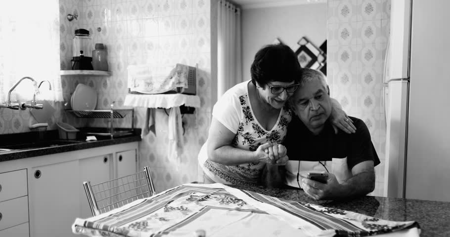 Struggling senior husband suffering from mental illness while elderly wife comforts and soothes partner during crisis of old age in kitchen scene in dramatic black and white