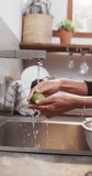 Vertical video of hands of biracial woman rinsing vegetables in kitchen, slow motion. Lifestyle, food, cooking, healthy lifestyle and domestic lifestyle, unaltered.