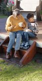 Vertical video of biracial grandmother and grandson sitting on terrace and holding mugs, slow motion. Family, togetherness and lifestyle, unaltered.