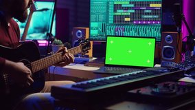 Musician beginner learning accords to play acoustic guitar by watching tutorials and online lessons, laptop shows greenscreen chromakey. Artist practicing guitar skills in home studio. Camera A.